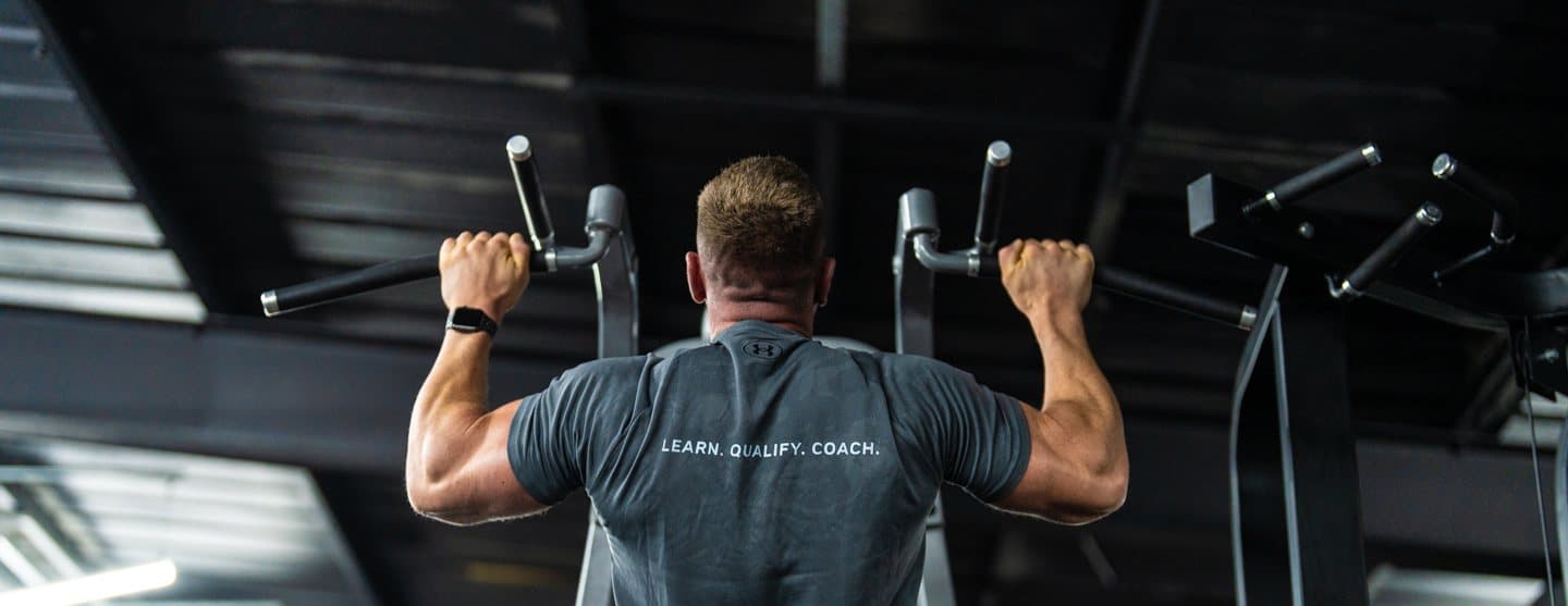 Clinton Coffey pulling down on a lat machine, LEARN. QUALIFY. COACH. across the back of his shirt
